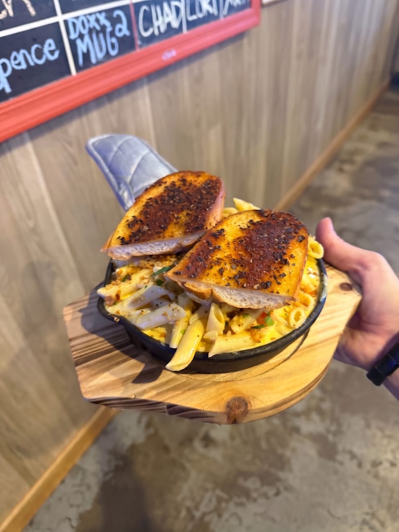 Hand holding a cast iron skillet of penne pasta with garlic bread on a wooden board.
