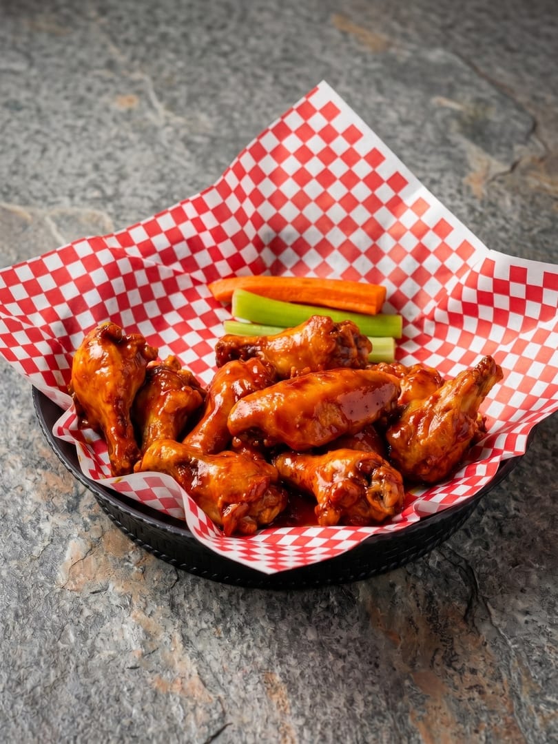 A basket of saucy chicken wings with celery and carrot sticks on red and white checkered paper.