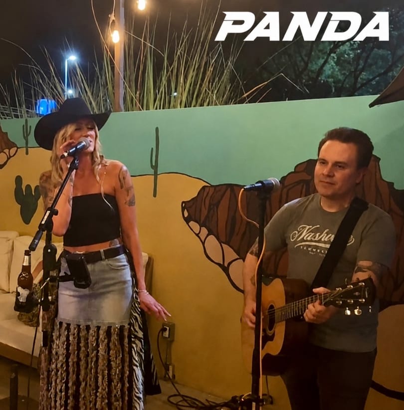 Woman in cowboy hat sings with a man playing guitar against a desert mural at night.