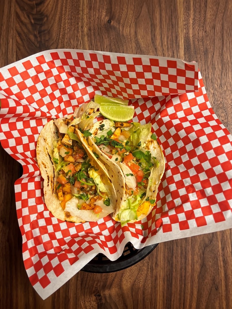 Two chicken tacos with lettuce, salsa, and lime wedges in a red and white checkered basket.