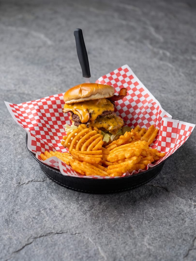 Double cheeseburger with bacon, melted cheese, and waffle fries in a basket with red checkered paper.