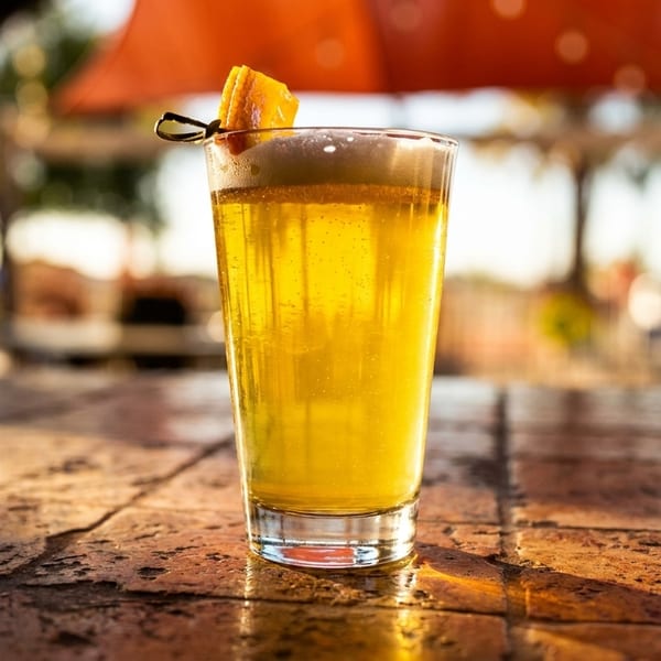 A tall glass of golden beer with a foamy head and orange garnish on a textured bar top.