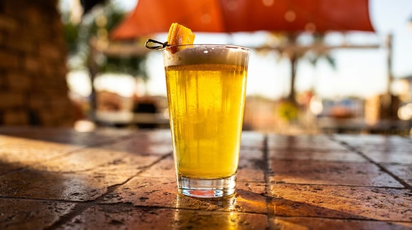 A tall glass of golden beer with a foamy head and orange garnish on a textured bar top.