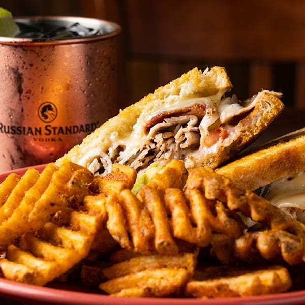 A hearty sandwich with melted cheese and waffle fries on a red plate, next to a Moscow Mule.