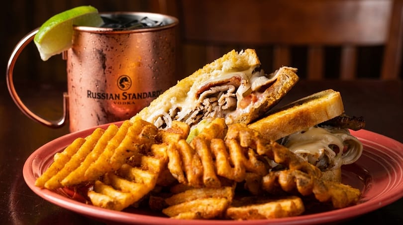 A hearty sandwich with melted cheese and waffle fries on a red plate, next to a Moscow Mule.