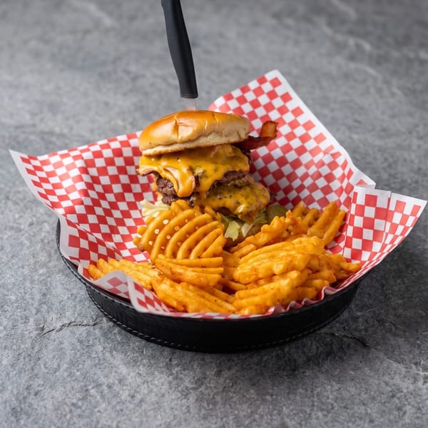 Double cheeseburger with bacon, melted cheese, and waffle fries in a basket with red checkered paper.