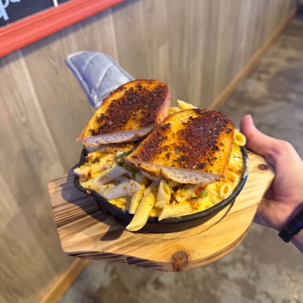 Hand holding a skillet of mac and cheese with garlic bread, on a wooden serving board.