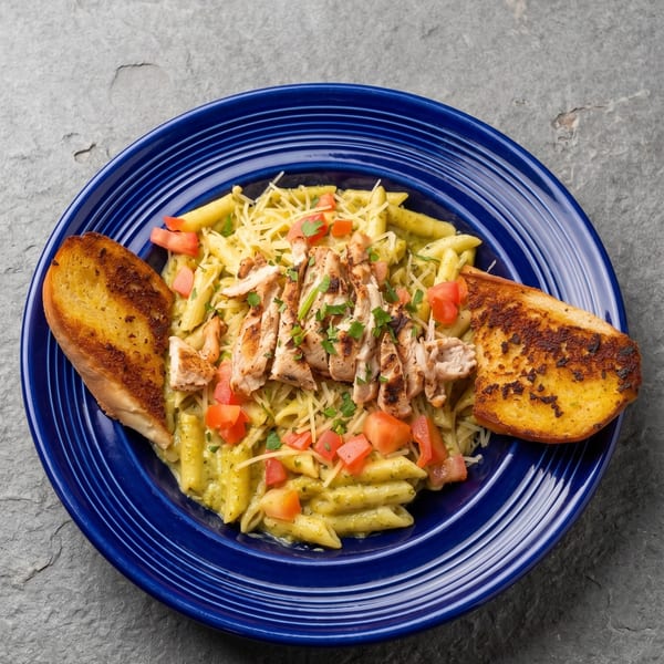 Penne pasta with pesto, grilled chicken, diced tomatoes, and garlic bread on a blue plate.