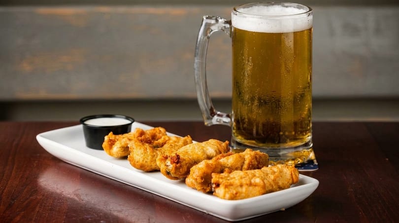 A mug of golden beer with foam next to a plate of fried appetizers and a small cup of dip.