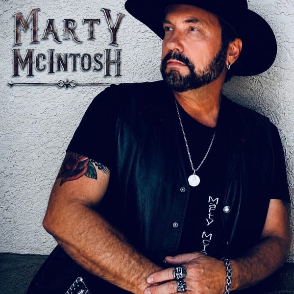 Musician Marty McIntosh in a black hat and vest, holding a guitar, with his name on the wall.