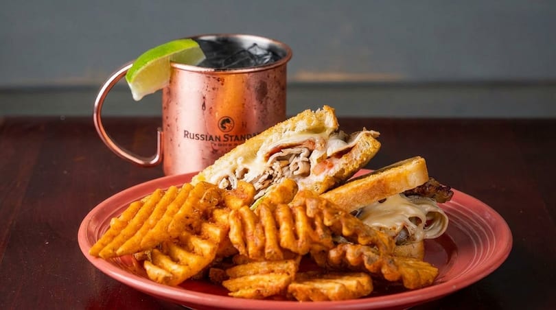 A grilled sandwich with waffle fries on a red plate, next to a copper mug with a lime.