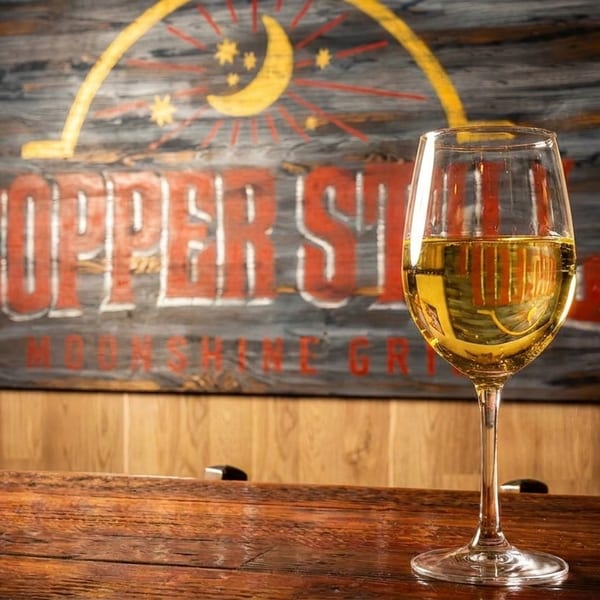 A glass of white wine on a wooden bar, with a "Copper Still Moonshine Grill" sign in the background.