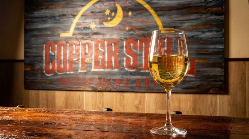 A glass of white wine on a wooden bar, with a "Copper Still Moonshine Grill" sign in the background.