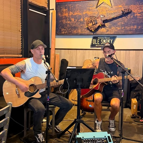 Two men perform live music, playing acoustic guitars and singing in a casual venue.