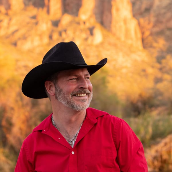 Smiling man in a black cowboy hat and red shirt against a golden desert mountain background.