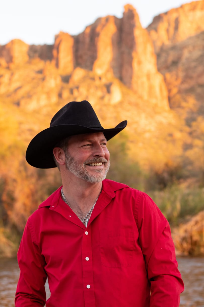 Smiling man in a black cowboy hat and red shirt against a golden desert mountain background.