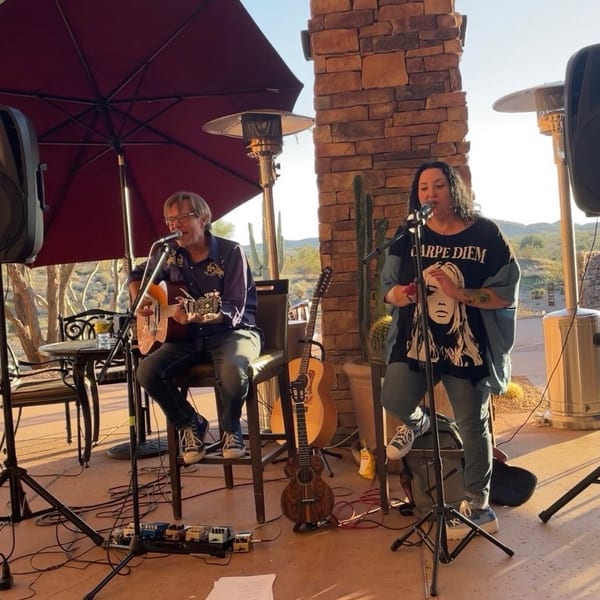 A man plays guitar and a woman sings on an outdoor patio with desert views.