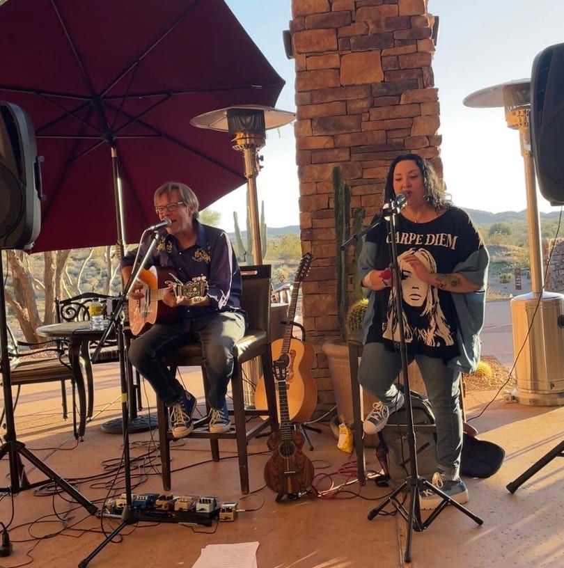 A man plays guitar and a woman sings on an outdoor patio with desert views.