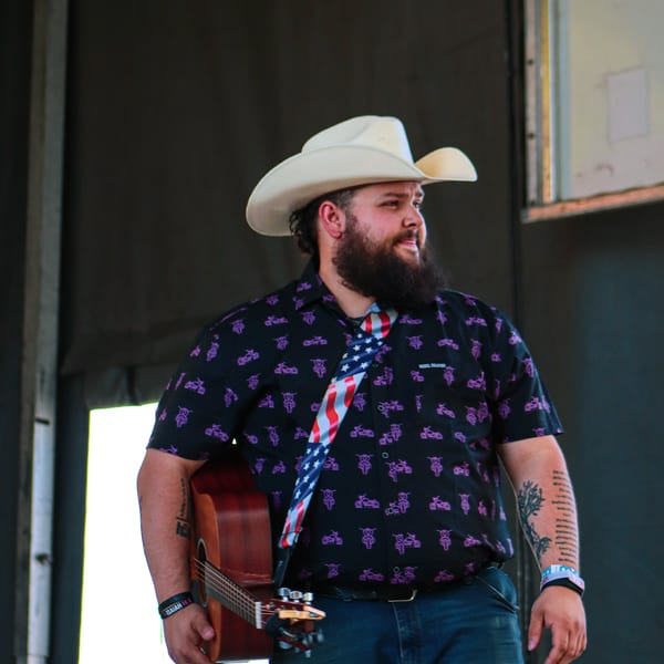 Musician in a cowboy hat and patterned shirt, holding an acoustic guitar with an American flag strap.
