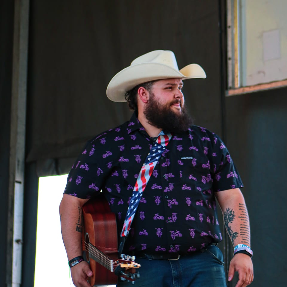 Musician in a cowboy hat and patterned shirt, holding an acoustic guitar with an American flag strap.