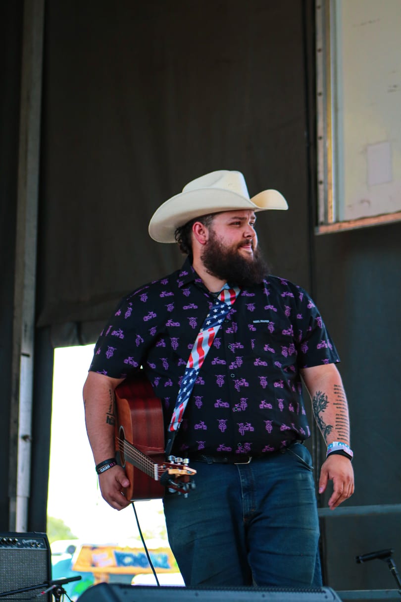 Musician in a cowboy hat and patterned shirt, holding an acoustic guitar with an American flag strap.